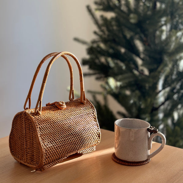 A wicker handbag with a guava-shaped design rests on a wooden surface next to a small cup, evoking a holiday atmosphere with a Christmas tree visible in the background. The bag features a distinctive bamboo-like pattern and a handle made from the same material.