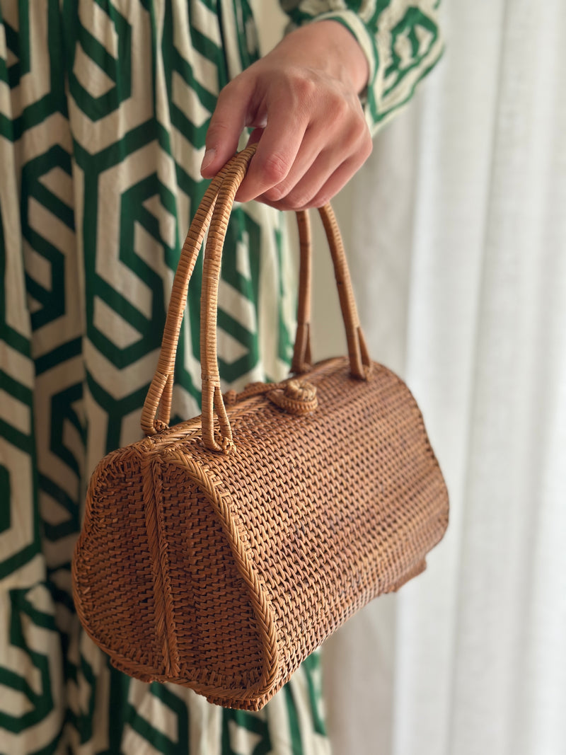 A person holding a woven rattan handbag with a unique guava-shaped design, featuring a light brown color and textured weave pattern. The bag has a sturdy handle made from natural rattan or similar fiber, resting on the surface of a blurred indoor setting with a green and white patterned wall.