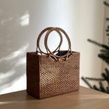 A woven rattan handbag, Bali Rattan Handbag - Hailey, rests on a wooden surface. The bag features two round Ata grass handles, adding elegance to its design. A soft Batik cotton lining ensures secure storage within the drawstring closure.