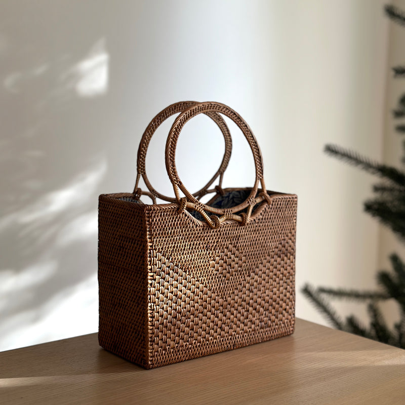 A woven rattan handbag, Bali Rattan Handbag - Hailey, rests on a wooden surface. The bag features two round Ata grass handles, adding elegance to its design. A soft Batik cotton lining ensures secure storage within the drawstring closure.