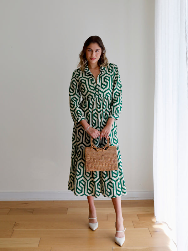 A woman stands near white curtains, holding a Bali Rattan Handbag - Hailey. The handbag features a smooth, polished rattan finish with a round handle made from Ata grass. A soft Batik lining and drawstring closure add to its secure storage capabilities.
