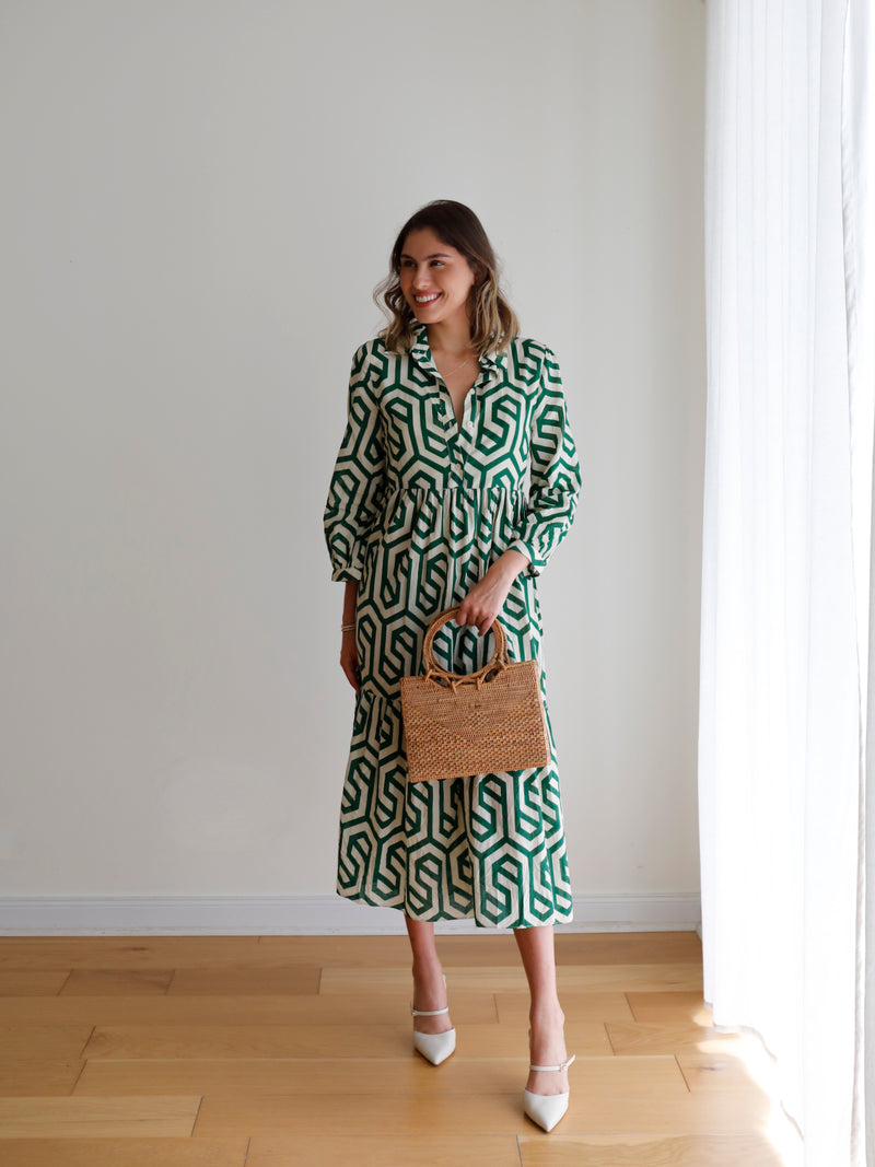 A woman stands indoors, wearing a long-sleeved patterned dress, holding a small rattan handbag with a round handle. The bag is made of rattan and features a smooth, polished finish.