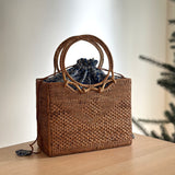 A woven rattan handbag, Bali Rattan Handbag - Hailey, sits on a surface adorned with a blue ribbon tied in a bow at the top. The bag features a round handle made of rattan and is lined with fabric.