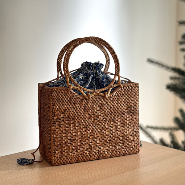 A woven rattan handbag, Bali Rattan Handbag - Hailey, sits on a surface adorned with a blue ribbon tied in a bow at the top. The bag features a round handle made of rattan and is lined with fabric.