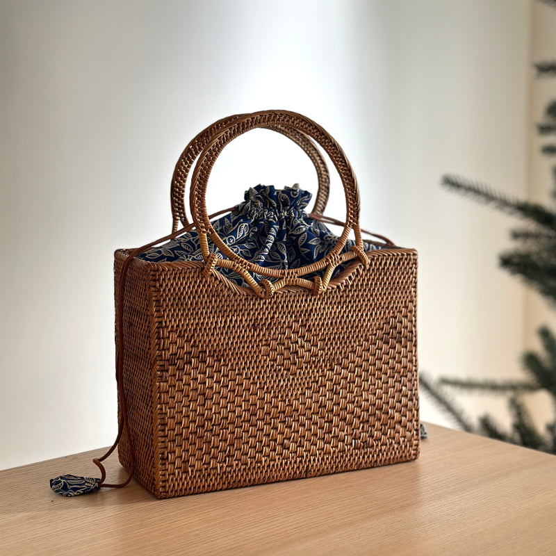 A woven rattan handbag, Bali Rattan Handbag - Hailey, sits on a surface adorned with a blue ribbon tied in a bow at the top. The bag features a round handle made of rattan and is lined with fabric.