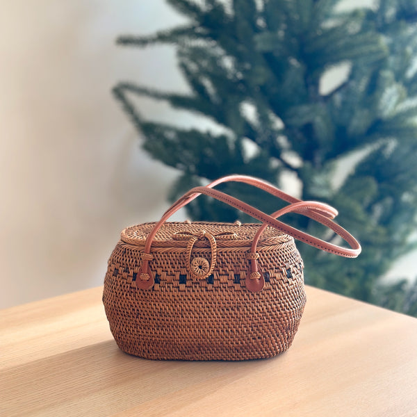 A woven rattan handbag, the Julia, rests on a wooden surface. The oval-shaped bag features a smooth, polished finish with a snag-free design. A genuine leather handle is secured by a bowtie clasp with secure snaps for closure. Lined with Batik cotton, the bag exudes a festive and cozy aesthetic.