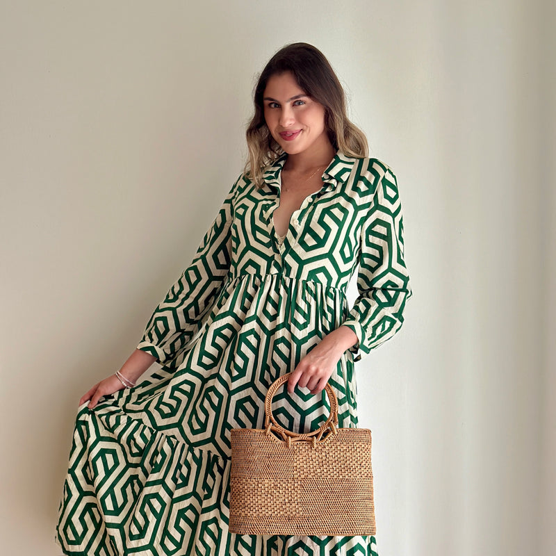 A woman stands in front of a white wall, wearing a green and white patterned jumpsuit with a belted waist. She holds a woven basket made from rattan or natural material, posing with one hand on her hip. The product, Bali Rattan Handbag - Kelsey, is the focus of the image.