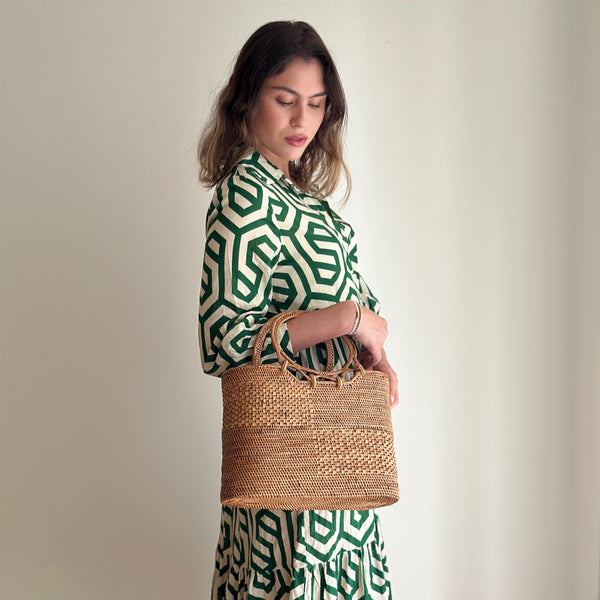 A woman stands against a plain background, wearing a long green dress with a geometric pattern. She holds a woven handbag with a textured surface, featuring a circular handle design that complements the dress's color. The bag appears to be made of rattan, with a natural material used for the handles.