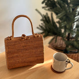 A woven rattan handbag, Bali Rattan Handbag - Kenia, rests on a wooden surface, positioned in front of a Christmas tree. The bag's natural texture and craftsmanship are highlighted, emphasizing its eco-friendly nature and unique design elements.