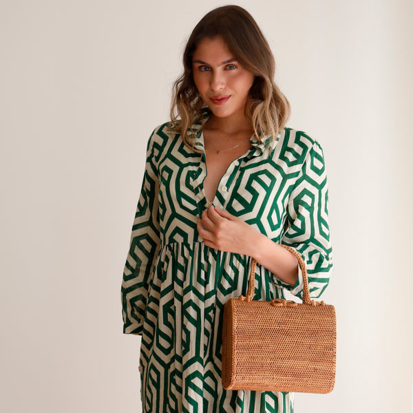 A woman stands in front of a white background, wearing a green and white patterned jumpsuit, holding a woven rattan handbag featuring a geometric design on the side. The bag has an envelope style with a short handle made from Ata grass, crafted using natural materials like bamboo or rattan.