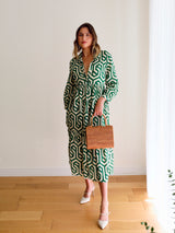 A woman stands against a white wall wearing a green and white patterned jumpsuit, holding the Bali Rattan Envelope Clutch. The clutch features a geometric design on its surface made from natural rattan material with a textured finish.