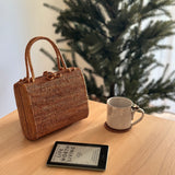 A woven rattan handbag, Bali Rattan Handbag - Kenia, sits on a wooden surface next to a small white mug. The handbag features a textured appearance with a unique design pattern or texture. Its spacious interior can hold daily essentials like a phone, lipstick, and credit cards.