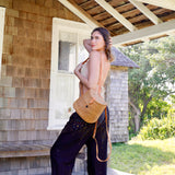A woman stands near a wooden structure wearing a sleeveless top, holding a Bali Rattan Mini Backpack made of wicker and rattan materials. The bag has adjustable leather straps and a batik lining, positioned on the right side of the frame.