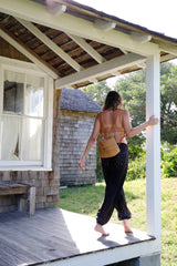 A person stands on a porch, holding a wooden railing with one hand. In their other hand is the Bali Rattan Mini Backpack, made from eco-friendly rattan and real leather straps. The backpack's sleek arch shape and secure snap closure are visible, along with its unique batik lining.