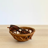 A pair of tortoiseshell sunglasses resting in a small, round handmade Bali rattan basket woven from ata grass. The Bali rattan basket sits on a light wood table, shown as a piece of unique home decor handmade in Bali.