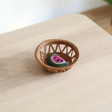A whimsical felt watermelon keychain sits in a small, round handmade Bali rattan basket woven from ata grass. This unique home decor rattan basket, handmade in Bali, is placed on a light wood table to hold small trinkets.