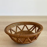 A single, small handmade Bali rattan basket, woven from ata grass, sits on a light wood table. The close-up shot highlights its intricate open-weave lattice, showcasing it as a piece of unique home decor handmade in Bali.
