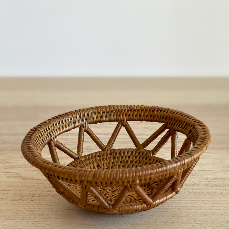 A single, small handmade Bali rattan basket, woven from ata grass, sits on a light wood table. The close-up shot highlights its intricate open-weave lattice, showcasing it as a piece of unique home decor handmade in Bali.