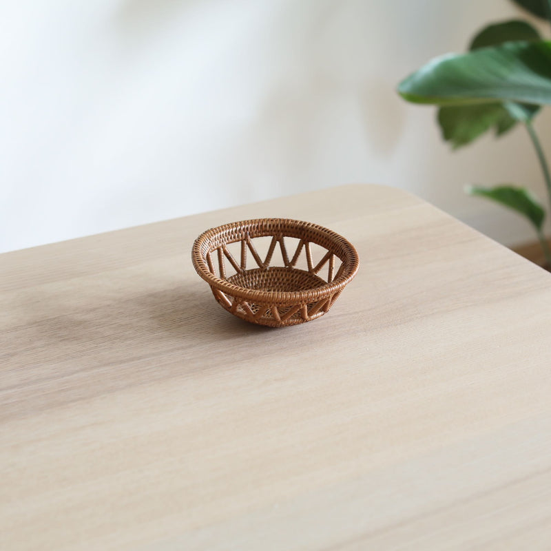 A small, handmade Bali rattan basket woven from ata grass, sitting on a light wood table in a home setting. This unique home decor piece, handmade in Bali, is set against a white wall with a soft-focus green plant in the background, creating a serene, natural feel.