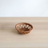 A small, round, honey-brown Bali rattan basket, handmade in Bali from ata grass, sits centered on a light wood table. Its unique home decor design features a solid woven base and sides with an airy, open-lattice pattern of triangles, set against a plain white wall.