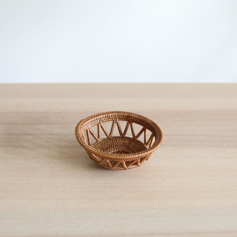 A small, round, honey-brown Bali rattan basket, handmade in Bali from ata grass, sits centered on a light wood table. Its unique home decor design features a solid woven base and sides with an airy, open-lattice pattern of triangles, set against a plain white wall.