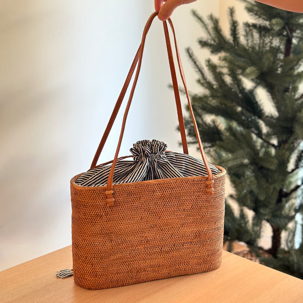 The Bali Rattan Shoulder Bag, a beautifully handwoven tote made from natural Ata grass, is held by its slim leather straps. Resting on a wooden table, the bag showcases its practical and stylish design, featuring a blue and white striped fabric lining that cinches closed with a secure drawstring top. In the background, a small evergreen tree adds a touch of natural ambiance, highlighting the bag's versatile, year-round appeal.