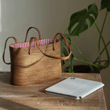 A rectangular, handwoven rattan tote bag rests on a rustic wooden table. The golden-brown bag has thin tan leather shoulder straps and an open top revealing a blue and white striped fabric lining. Beside it, a silver tablet with a white cover lies on the table. To the right, blurred green monstera leaves provide a natural backdrop, with soft light from the left casting a shadow on the wall.