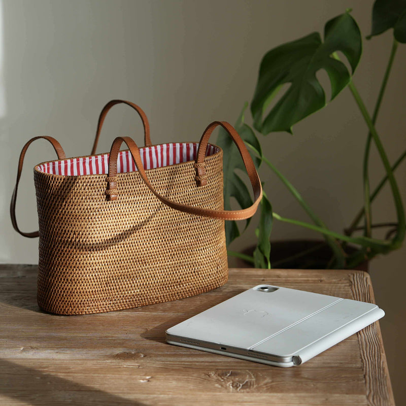 A rectangular, handwoven rattan tote bag rests on a rustic wooden table. The golden-brown bag has thin tan leather shoulder straps and an open top revealing a blue and white striped fabric lining. Beside it, a silver tablet with a white cover lies on the table. To the right, blurred green monstera leaves provide a natural backdrop, with soft light from the left casting a shadow on the wall.
