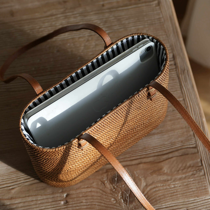 A high-angle, close-up photograph shows a handwoven, rectangular rattan tote bag resting on a rustic wooden table. The bag is filled with a silver tablet, demonstrating its size and capacity. The interior of the golden-brown bag is lined with a black-and-white striped fabric, and its thin, tan leather shoulder straps rest on the table. Strong natural light from the left highlights the intricate weave of the rattan and creates a bright glare on the tablet's screen.