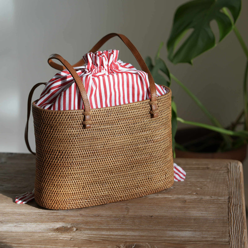 A rectangular, handwoven rattan tote bag with a golden-brown finish sits on a rustic wooden table. The bag is lit from the left by natural sunlight, highlighting its intricate texture. It features brown leather handles and a vibrant red-and-white vertically striped fabric lining that is pulled closed with a drawstring, creating a ruffled top that peeks out of the bag. A small piece of the striped drawstring hangs over the left edge of the bag. In the background, a large green monstera leaf is visible.
