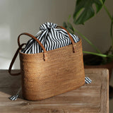 A rectangular, handwoven rattan tote bag with a golden-brown finish sits on a rustic wooden table. The bag is lit from the left by natural sunlight, highlighting its intricate weave. It features brown leather handles and a black-and-white vertically striped fabric lining that is pulled closed with a white drawstring, creating a gathered top that peeks out of the bag. A small, tassel-like end of the drawstring rests on the table. In the background, a large green monstera leaf is visible against a plain wall.
