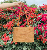 A woven Bali Rattan Shoulder Bag is held against vibrant red flowers, set amidst lush greenery. The bag is handwoven from natural rattan material with a textured surface, indicating handmade craftsmanship.