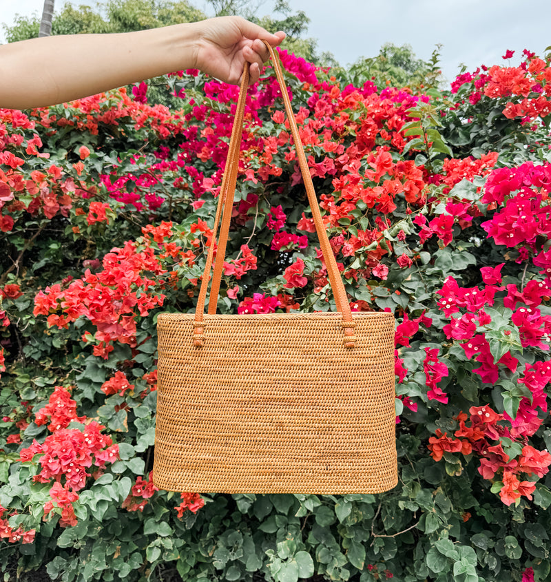 A woven Bali Rattan Shoulder Bag is held against vibrant red flowers, set amidst lush greenery. The bag is handwoven from natural rattan material with a textured surface, indicating handmade craftsmanship.