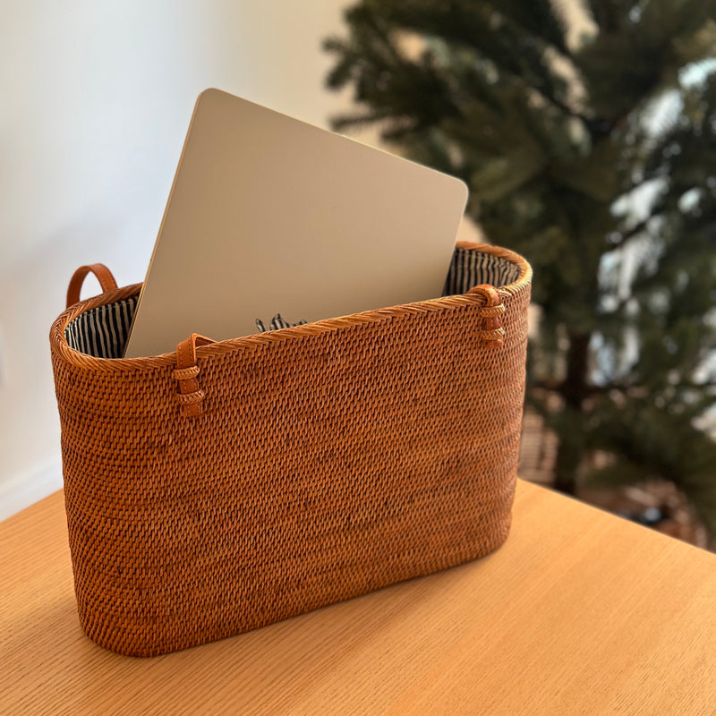 A Bali Rattan Shoulder Bag rests on a wooden surface, open to reveal its interior featuring a drawstring pouch. A laptop is visible inside the bag, suggesting its use for carrying personal items or work essentials. The background features a Christmas tree and part of a person's arm, conveying a cozy home setting during the holiday season.