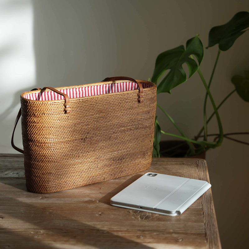 The handcrafted Bali Rattan Shoulder Bag sits elegantly on a rustic wooden table, bathed in natural sunlight. Its sturdy, rectangular shape and intricate weave highlight its artisanal quality, while the vibrant red-and-white striped interior lining adds a playful contrast. Pictured next to a tablet, the spacious tote proves both stylish and functional, perfect for carrying daily essentials. In the background, a monstera plant enhances the scene's natural, relaxed aesthetic.