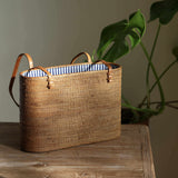This beautifully handcrafted Bali Rattan Shoulder Bag, made from natural woven Ata grass, rests on a rustic wooden table in a sunlit room. Its classic tote design is elevated by rich, brown leather handles and a charming blue-and-white striped interior lining. Ethically handmade in Bali, this eco-friendly bag combines timeless style with sustainable craftsmanship, making it a perfect accessory for any occasion. A monstera plant in the background enhances its natural, artisanal appeal.