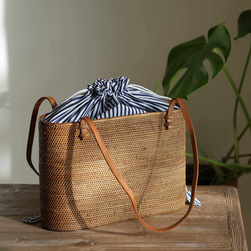 A rectangular, handwoven rattan tote bag sits on a rustic wooden table in a warmly lit room. The bag has thin, brown leather shoulder straps attached to the top rim. Peeking out from the top is a blue and white striped fabric lining, which is gathered and tied shut with a white drawstring. Sunlight from the left highlights the texture of the weave and casts a soft shadow behind it. In the blurred background, the large, dark green leaves of a monstera plant are visible.