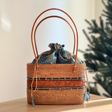 A woven rattan tote bag rests on a surface, showcasing its brown color with a patterned design. The bag features a real leather handle and drawstring closure with a batik fabric lining, adding a touch of elegance and sophistication.