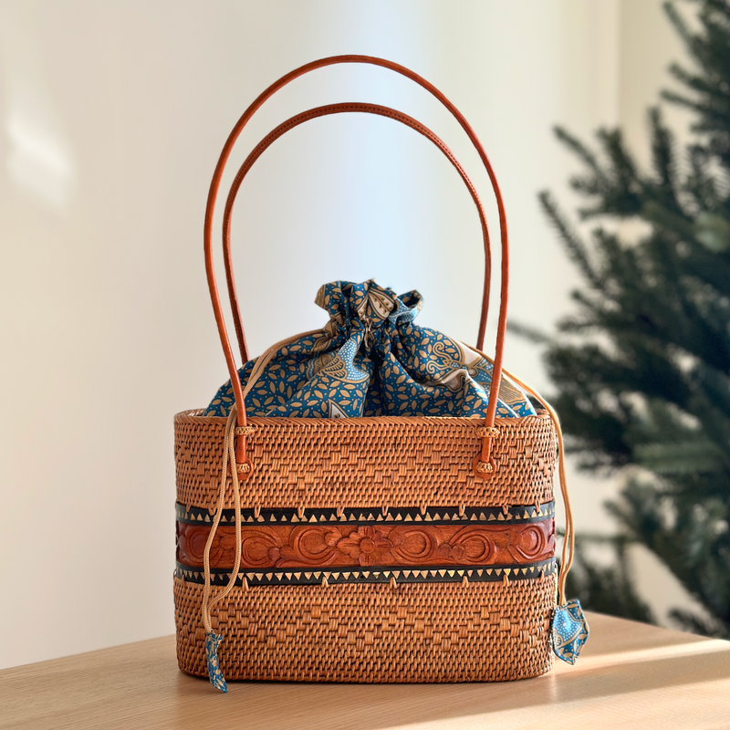 A woven rattan tote bag rests on a surface, showcasing its brown color with a patterned design. The bag features a real leather handle and drawstring closure with a batik fabric lining, adding a touch of elegance and sophistication.