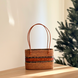 A Bali Rattan Tote handcrafted with intricate wood carvings rests on a wooden surface amidst a festive holiday backdrop. The tote features a brown leather handle, smooth rattan finish, and a Batik cotton drawstring pouch inside. Its relaxed silhouette and polished texture evoke warmth and comfort.