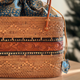 A Bali Rattan Tote – Handcrafted Wood Carving Edition, resting on a wooden table, features a real leather handle and a Batik lining drawstring pouch. The tote showcases an elegant and durable design with visible wooden texture, conveying comfort and luxury.