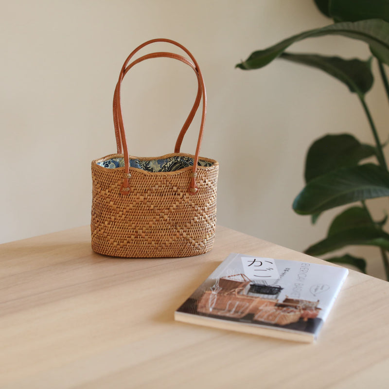 A woven rattan handbag, Bali Rattan Wave Handbag - Kauai, is placed on a wooden surface next to a book. The background features a plant, creating a relaxed setting within a home or cozy cafe. The oval-shaped bag has a top handle and luxurious leather handles, with a Batik cotton lining and drawstring pouch.