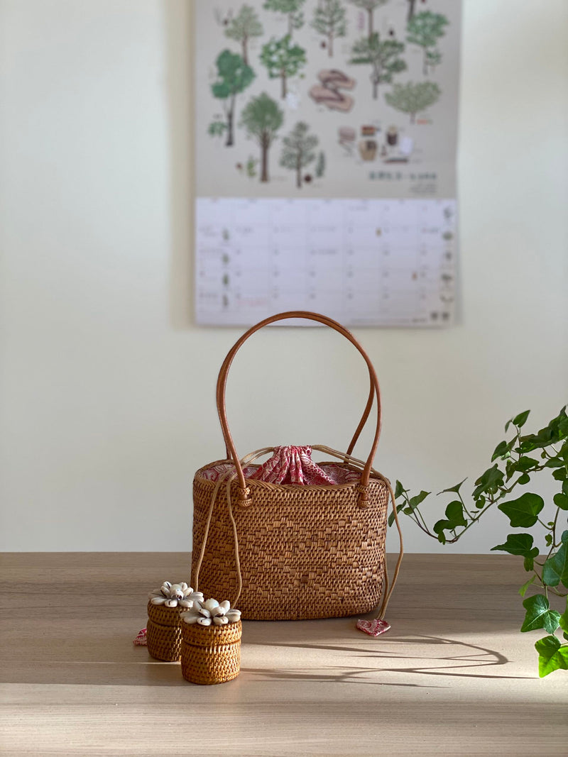 A woven handbag with a distinctive oval shape, made from rattan material with a textured weave, rests on a wooden surface. The bag features a handle on top, drawstring closure, and is lined with soft Batik cotton fabric.The bag is presented in a casual, homey setting.