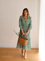A woman stands against a white wall, holding a woven rattan handbag in her right hand. The bag features a geometric pattern and appears to be made of natural materials with a textured surface. It matches the design on her long-sleeved dress.