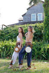 Ava Crossbody Bag by Bali, resting on one woman's shoulder as she stands outdoors in a sunny environment. The bag has a round shape, rattan handle adorned with seashells, and appears made from natural materials. A bohemian-style outfit complements the bag's vibrant summer vibe.