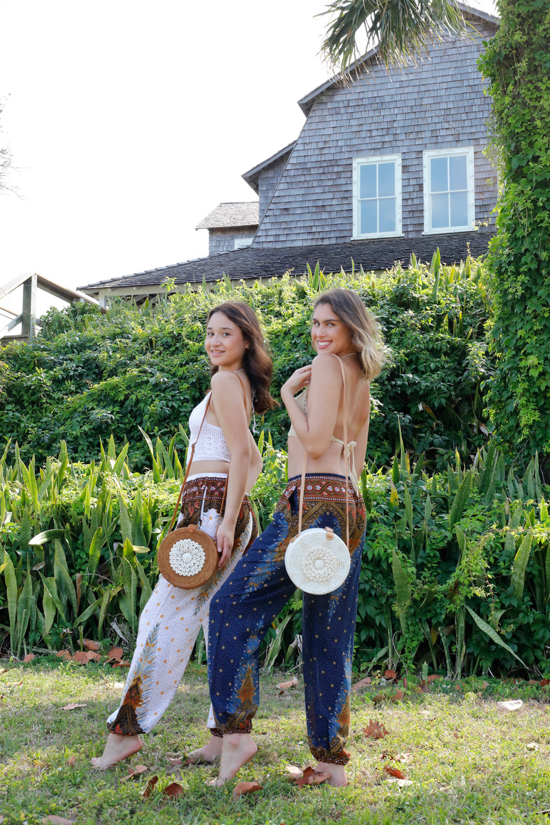 Ava Crossbody Bag by Bali, resting on one woman's shoulder as she stands outdoors in a sunny environment. The bag has a round shape, rattan handle adorned with seashells, and appears made from natural materials. A bohemian-style outfit complements the bag's vibrant summer vibe.