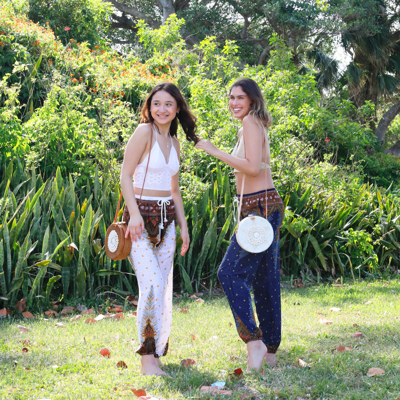 A seashell crossbody bag, made from natural rattan with real seashells and rattan accents, is being showcased by two women enjoying their time outdoors in a park-like setting. The woman on the right is holding her bag in front of her, while the other bag is hanging from the woman's shoulder. The bags feature a bohemian touch with real seashells and rattan accents.