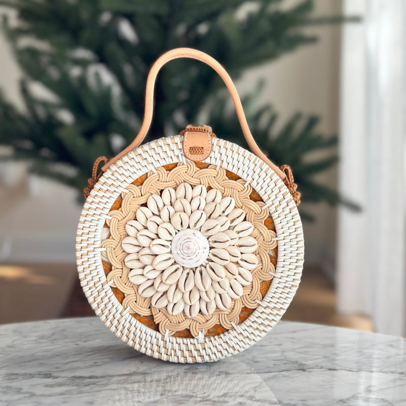 A woven rattan crossbody bag with a distinctive seashell pattern on its surface rests on a table, surrounded by a cozy indoor setting featuring a plant and a portion of a Christmas tree. The bag's natural materials and adjustable strap suggest it can be carried in multiple ways, while the circular shape with floral pattern gives it a bohemian aesthetic.