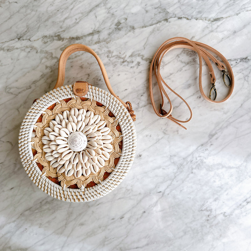 A woven rattan crossbody bag, round in shape and featuring a floral pattern, rests on a marble surface next to a pair of sunglasses with a similar color scheme. The bag has a snap closure, an adjustable strap, and a leather handle, evoking a relaxed and leisurely mood reminiscent of a beach or resort location.