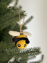 A cheerful felted wool bee ornament hangs from a branch of an evergreen Christmas tree. This handmade ornament features a round yellow and black striped body with small, delicate white wings, suspended by a jute string, adding a touch of handcrafted charm.